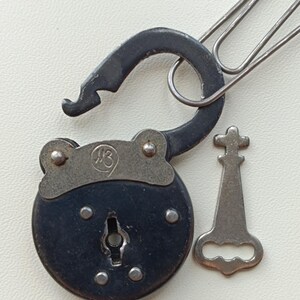 May include: An antique black padlock with a silver key and a paperclip. The lock is open, revealing the keyhole. The key is silver and has a cross-shaped top. The lock has a circular body with rivets and a curved shackle.