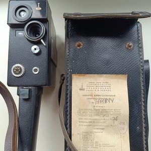 May include: A vintage black film camera with a leather carrying case. The camera has a lens, dials, and a handle. The case is dark blue with a strap and a document inside. The camera and case are placed on a light-colored surface.