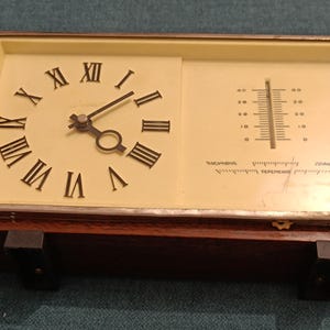 May include: A vintage wooden clock with a white face and Roman numeral hour markers. The clock also has a thermometer with a Fahrenheit scale. The clock is in a wooden frame with a dark brown finish.