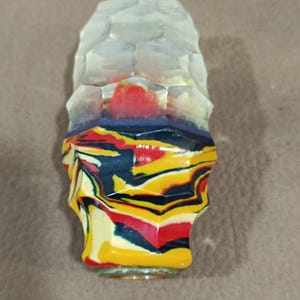 Puede incluir: Un tapón de botella decorativo con una parte superior transparente y facetada y una base colorida. La base presenta un patrón arremolinado de amarillo, rojo, azul y negro. El tapón está diseñado para adaptarse a una botella estándar.