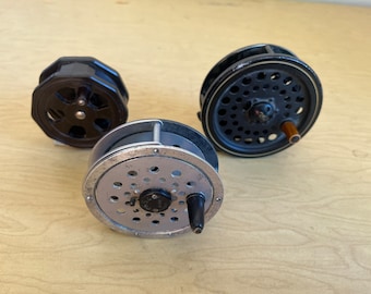 May include: Three vintage fly fishing reels. The reels are black and silver, with various designs and handle styles. The reels are arranged on a light-colored wooden surface.