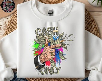 May include: White sweatshirt featuring a graphic of a hand holding cash, with the words "CASH ONLY". The design includes colourful paint splatters and dollar bills. The hand has tattoos and gold rings.