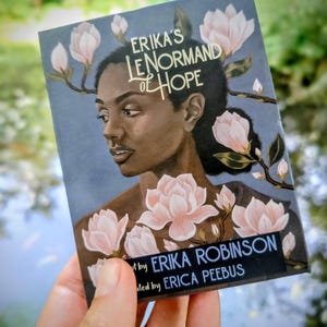 May include: A rectangular deck of cards titled Erika's Lenormand of Hope, featuring an illustration of a person surrounded by pink magnolia flowers against a dark background. The text reads Erika's Lenormand of Hope, created by Erika Robinson, illustrated by Erica Peebus.