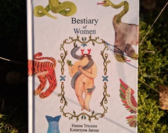 May include: A book titled "Bestiary of Women" with illustrations of a tiger, snakes, and a nude figure. The cover art features a woman with antlers and a snake wrapped around her. The book is set against a backdrop of green foliage.
