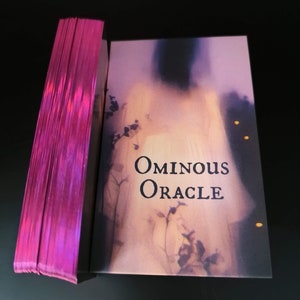 May include: A deck of cards titled "Ominous Oracle" with a pink iridescent edge. The card box features a blurred image of a figure and the title in black font. The cards are stacked next to the box on a black surface.
