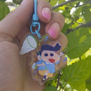 Può includere: Un portachiavi in plastica trasparente con un personaggio dei cartoni animati che tiene una rete per catturare farfalle. Il personaggio ha i capelli neri, una camicia blu e indossa un vestito giallo e blu. Lo sfondo è pieno di farfalle e stelle.