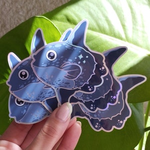 Puede incluir: Tres pegatinas de peces azul y negro iridiscentes con acabado holográfico. Los peces están apilados uno encima del otro y tienen una apariencia de dibujos animados.