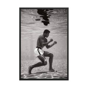 Puede incluir: Fotografía en blanco y negro de un boxeador bajo el agua, enmarcada en negro. El boxeador está en posición de combate, vistiendo pantalones cortos blancos con una raya negra y un cinturón negro. La superficie del agua refleja la imagen del boxeador.