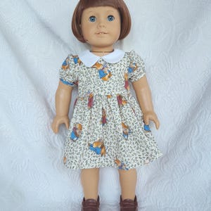 May include: A doll wearing a cream-colored dress with a Peter Rabbit print. The dress has a white collar and short sleeves with blue trim. The doll has brown shoes and a short brown bob hairstyle.