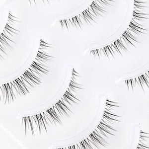 Può includere: Primo piano di diversi set di ciglia finte. Ogni set è nero e ha una forma curva, progettata per adattarsi alla palpebra naturale. Le ciglia sono lunghe e vaporose, creando un aspetto pieno e voluminoso. Sono confezionate in un contenitore di plastica trasparente.