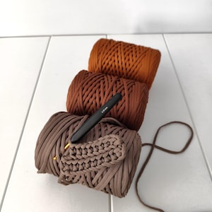 May include: Three cylindrical spools of cord yarn in shades of rust, medium brown, and light taupe. A black crochet hook with a 4.50 mm tip is resting on the taupe spool, which features a small section of brown crocheted fabric attached to it.