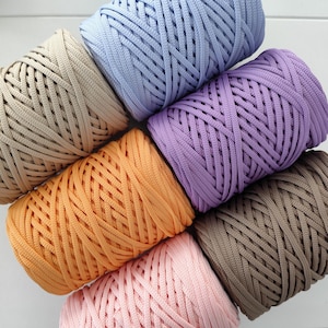 Puede incluir: Primer plano de varios carretes de hilo de macramé colorido en tonos beige, azul claro, naranja, morado, marrón y rosa. Los hilos están bien enrollados, mostrando la textura tejida. Ideal para proyectos de manualidades.