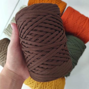 May include: A cylindrical spool of dark brown macrame cord featuring a braided texture. The cord is wound tightly in a continuous pattern. Several other spools of cord in various earth tones, including orange, yellow, and olive green, are visible.