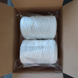 Op de afbeelding: Twee witte spoelen koord zijn verpakt in doorzichtig plastic in een kartonnen doos. Het koord lijkt gemaakt van een geweven materiaal. De doos is open en onthult de inhoud.
