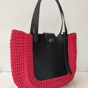 Op de afbeelding: Handgemaakte tote bag met een zwarte leren body en zwarte leren bandjes. De tas heeft een levendige, felroze gehaakte rand. De tas heeft een zilverkleurige sluiting en staat tegen een witte achtergrond.
