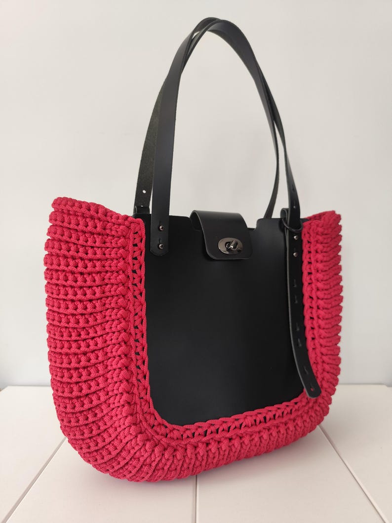 Op de afbeelding: Handgemaakte tote bag met een zwarte leren body en zwarte leren bandjes. De tas heeft een levendige, felroze gehaakte rand. De tas heeft een zilverkleurige sluiting en staat tegen een witte achtergrond.
