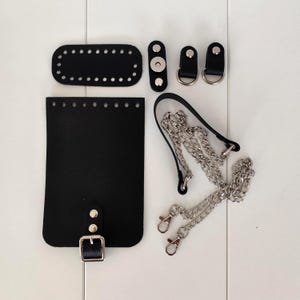 Op de afbeelding: Een zwarte leren tassenmaakset met diverse onderdelen. Inclusief een rechthoekige basis, een rechthoekig lichaam, twee D-ringen, een magneetsluiting, een gesp en een zilveren kettingriem met sluitingen. Alle onderdelen zijn zwart.