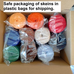 Op de afbeelding: Een kartonnen doos gevuld met strengen garen in verschillende kleuren, waaronder oranje, rood, blauw en groen. Elke streng is afzonderlijk verpakt in doorzichtig plastic. De tekst luidt: "Veilige verpakking van strengen in plastic zakken voor verzending."