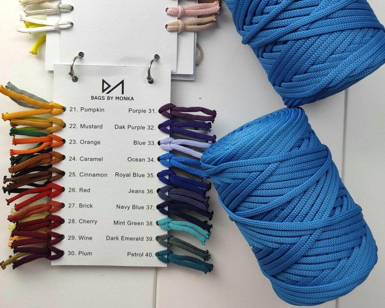 Op de afbeelding: Een bovenaanzicht toont een kleurenkaart en twee grote spoelen blauw koord. De kaart, met het label "BAGS BY MONKA", bevat verschillende koordkleuren, waaronder Pompoen, Mosterd en Rood. De spoelen koord zijn een levendige blauwe tint, perfect voor knutselen.