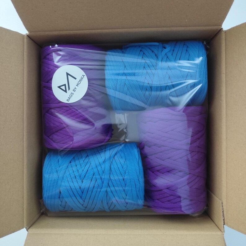 Op de afbeelding: Een kartonnen doos met vier spoelen gekleurd koord. Twee spoelen zijn helderblauw en twee zijn dieppaars. De spoelen zijn verpakt in doorzichtig plastic. De tekst "BAGS BY MONKA" is gedrukt op een witte ronde sticker.