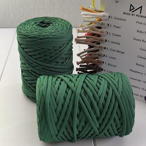 Op de afbeelding: Twee spoelen groen macram&eacute;koord worden getoond, &eacute;&eacute;n rechtop en &eacute;&eacute;n liggend. Een kleurenkaart met verschillende koordkleuren is zichtbaar op de achtergrond, met de tekst "BAGS BY MONKA" bovenaan. Het koord lijkt gemaakt van een geweven materiaal.