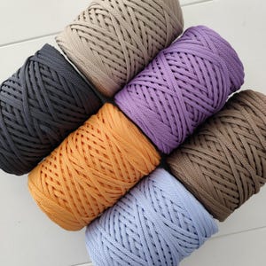 Op de afbeelding: Een bovenaanzicht toont zeven spoelen kleurrijk, dik, gevlochten garen. De garenkleuren zijn grijs, beige, paars, oranje, bruin en lichtblauw. De spoelen zijn diagonaal gerangschikt, wat de textuur en verscheidenheid van het garen benadrukt.