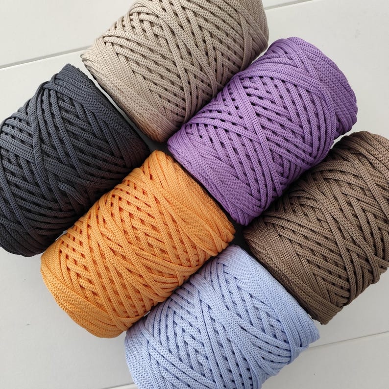 Op de afbeelding: Een bovenaanzicht toont zeven spoelen kleurrijk, dik, gevlochten garen. De garenkleuren zijn grijs, beige, paars, oranje, bruin en lichtblauw. De spoelen zijn diagonaal gerangschikt, wat de textuur en verscheidenheid van het garen benadrukt.