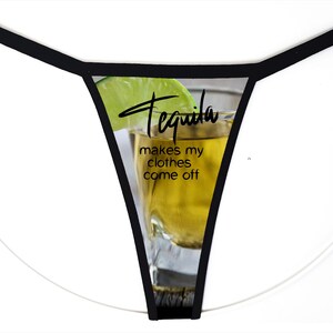 May include: A thong with black trim featuring a graphic print of a shot glass filled with amber liquid and a lime wedge. The text printed on the front reads, "Tequila makes my clothes come off" in a combination of script and sans-serif fonts.