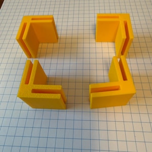 May include: Four bright yellow L-shaped corner brackets, each featuring a distinct slot along the inner edges. These rigid plastic components are designed with a right-angle geometry, providing a stable frame for connecting two perpendicular surfaces.