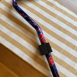 May include: A walking cane featuring a dark blue metallic finish with a vibrant floral pattern in shades of red, purple, and blue. The cane includes a black foam handle grip at the top and a black rectangular adjustment clamp on the shaft.