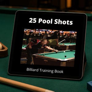 Puede incluir: Una tableta digital que muestra una portada con el texto 25 Pool Shots y Billiard Training Book, con una imagen de una persona jugando al billar. La tableta está colocada sobre una superficie de fieltro verde con bolas y un taco.