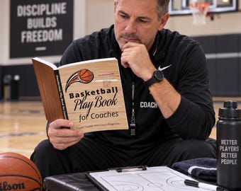 Op de afbeelding: Een softcover boek getiteld Basketball Play Book for Coaches met een houtnerf ontwerp op de kaft, voorzien van een basketbalafbeelding, een basketring en de tekst Player Tracker With Court Diagrams, opengehouden om de voorzijde te tonen.