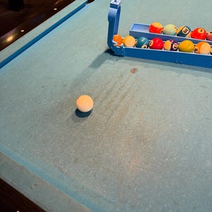 May include: A light blue pool table with a white cue ball resting on the felt surface. A light blue plastic ball rack holds several multi-colored numbered billiard balls, including red, yellow, blue, and orange spheres, positioned near the edge.