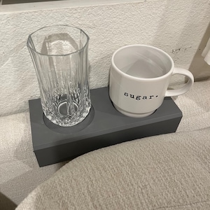 May include: A gray ceramic coaster with two indentations, one holding a clear glass and the other holding a white ceramic mug with the word "sugar" printed on it.