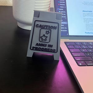 May include: A gray, 3D printed sign with black text that reads "CAUTION ANKI IN PROGRESS". The sign is shaped like a construction sign and has two stars in the middle of the text. The sign is sitting on a black surface in front of a laptop.