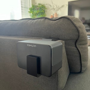 Può includere: Un altoparlante nero con la scritta "ULTIMEA" stampata su di esso è montato sul lato di un divano grigio.