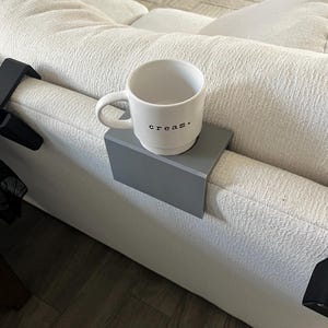 Puede incluir: Un portavasos gris sujeto al brazo de un sofá blanco. Una taza de cerámica blanca con la palabra "cream." impresa en ella está sentada en el portavasos.