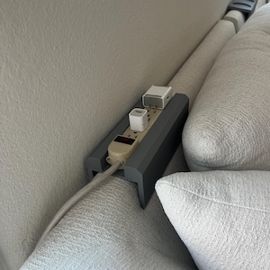 Könnte beinhalten: Eine graue Steckdosenleiste mit mehreren Steckdosen und zwei weißen Ladeadaptern. Die Steckdosenleiste ist an der Seite eines hellfarbigen Sofas montiert, mit einer weißen Wand im Hintergrund. Ein weißes Netzkabel ist eingesteckt.