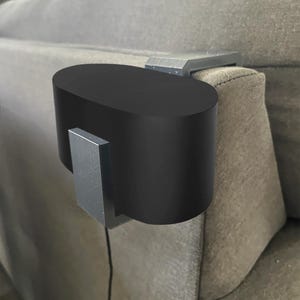 May include: A black, oval-shaped couch arm table with two silver-colored brackets. The table is designed to attach to the arm of a sofa. The product is made of solid materials.