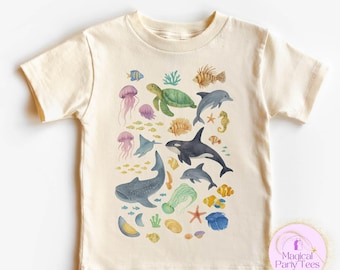 May include: Cream-colored t-shirt featuring a colorful watercolor design of various sea creatures. The design includes a sea turtle, dolphins, jellyfish, and a whale shark. The shirt has short sleeves and a crew neck.