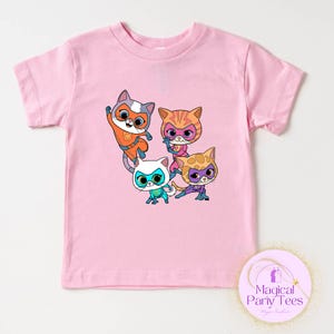 May include: Pink short-sleeved t-shirt featuring a graphic of four cartoon cats in superhero costumes. The cats are in various colors, including orange, pink, white, and yellow, with matching masks. The shirt has a round neckline.