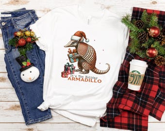 May include: White t-shirt featuring a festive armadillo wearing a Santa hat and holding gifts. The text "It's The Holiday Armadillo" is printed below the armadillo. The shirt is displayed with blue jeans, a snowman ornament, and holiday decor.