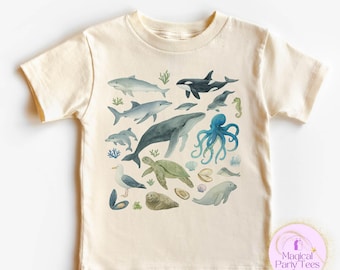 Può includere: T-shirt color crema con un'illustrazione ad acquerello di varie creature marine, tra cui balene, delfini e un polpo. Il design include un cavalluccio marino, una tartaruga e una foca. Il testo "Magical Party Tees" è visibile in basso a destra.