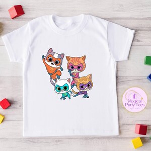May include: White t-shirt featuring a colorful graphic of four cartoon cat superheroes. The cats are in various poses and wearing masks and superhero outfits in orange, pink, blue, and purple. The shirt has a crew neck and short sleeves.