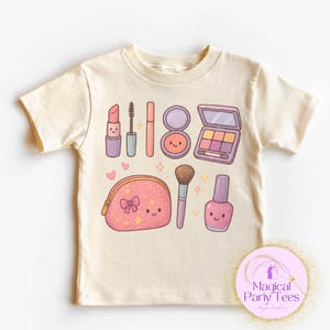 May include: Cream-colored t-shirt featuring a collection of cute makeup illustrations. The design includes a lipstick, mascara, blush, eyeshadow palette, makeup bag, brush, and nail polish. The shirt also has the text "Magical Party Tees".