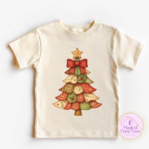 May include: A cream-coloured t-shirt with a Christmas tree design. The tree is made of patchwork shapes in red, green, and tan, decorated with a red bow and a gold star. The design has a rustic, festive aesthetic.