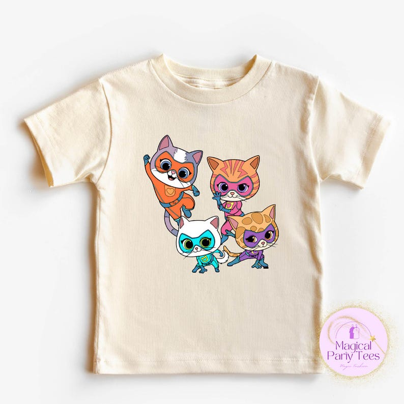 May include: Cream-colored short-sleeved t-shirt featuring a colorful graphic of four cartoon cats in superhero costumes. The cats are in various poses, with vibrant orange, pink, blue, and yellow accents. The shirt has a crew neck.