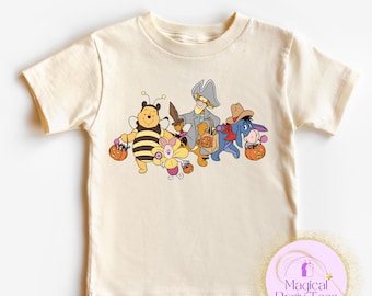 May include: A cream-coloured t-shirt with a cartoon illustration of Winnie the Pooh characters in Halloween costumes, each carrying a pumpkin-shaped candy bucket. The characters include Winnie the Pooh as a bee, Piglet, Tigger, Eeyore, and a pirate. The shirt has the text "Magical Party Tees".