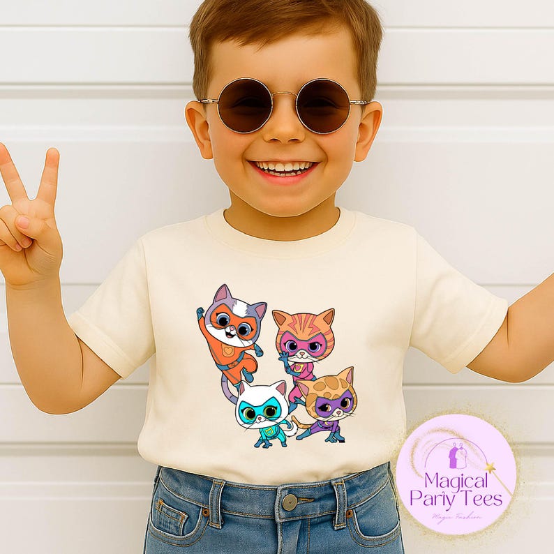 May include: Cream-colored t-shirt featuring a group of cartoon cat characters in superhero costumes. The cats are in various poses and colors, including orange, pink, blue, and purple. The shirt is worn by a child wearing sunglasses.