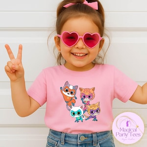 May include: A pink t-shirt featuring a cartoon graphic of four cat characters with superhero masks. The child in the image is wearing heart-shaped sunglasses and blue jeans. The t-shirt has the text "Magical Party Tees" on the bottom right.